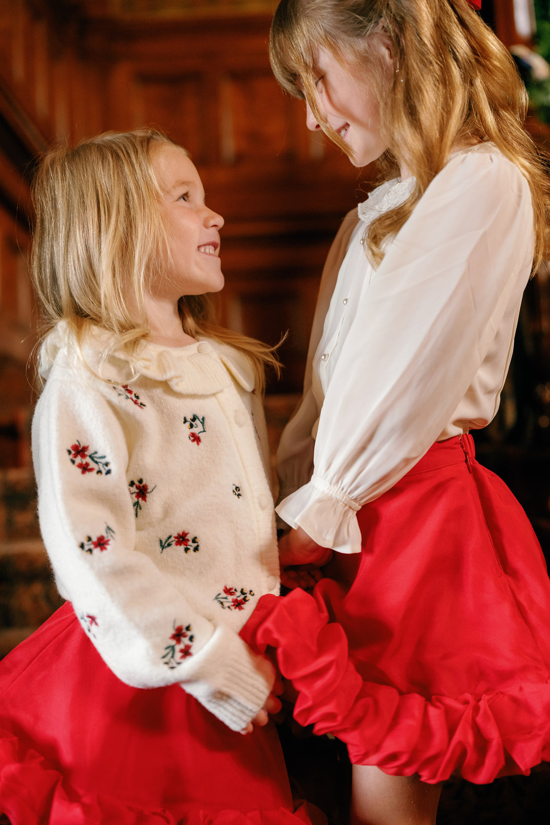 Two young girls with blonde hair, wearing white tops and the Ivy City Co Mini Kendy Skirt in Poinsettia, smile at each other indoors. One girl wears a floral-patterned sweater, and they appear happy as they hold their festive organza skirts and stand close together. Their matching holiday party outfits make the moment even more cheerful.
