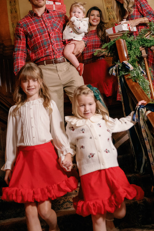 A family in festive red and white poses on a staircase adorned with greenery. At the front, two young girls hold hands, each wearing a charming girls’ A-line skirt—one features the Ivy City Co Mini Kendy Skirt in Poinsettia, its beautiful red organza making for the perfect holiday party outfit—while adults and a baby smile behind them, creating a joyful holiday scene.