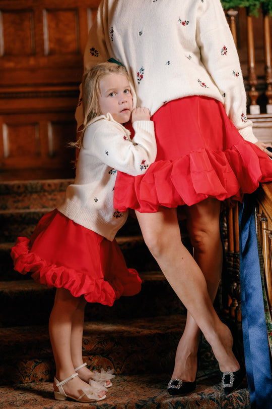 A young girl hugs an adult’s waist as they both wear matching white sweaters and Ivy City Co’s Mini Kendy Skirt in Poinsettia, creating a charming holiday party look. They stand on carpeted stairs beside a wooden railing.