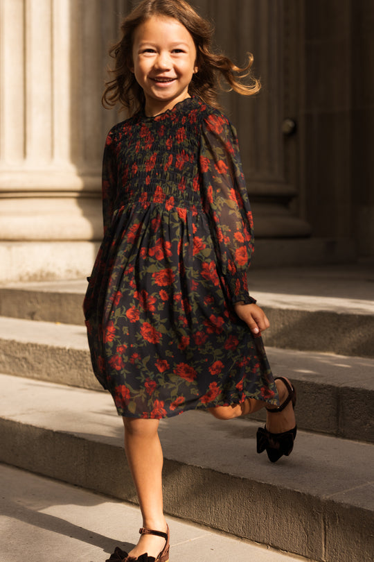 A young girl beams as she joyfully walks down stone steps, sunlight illuminating her and the grand columns behind her, dressed in the Ivy City Co Mini Linden Dress in Scarlet Blooms—a dark floral girls dress with charming balloon sleeves—paired perfectly with brown shoes.