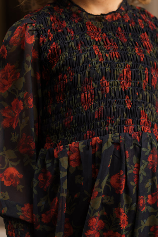 A person wearing the Ivy City Co Mini Linden Dress in Scarlet Blooms, featuring red floral patterns with green leaves, smocking on the bodice, and sheer long balloon sleeves.