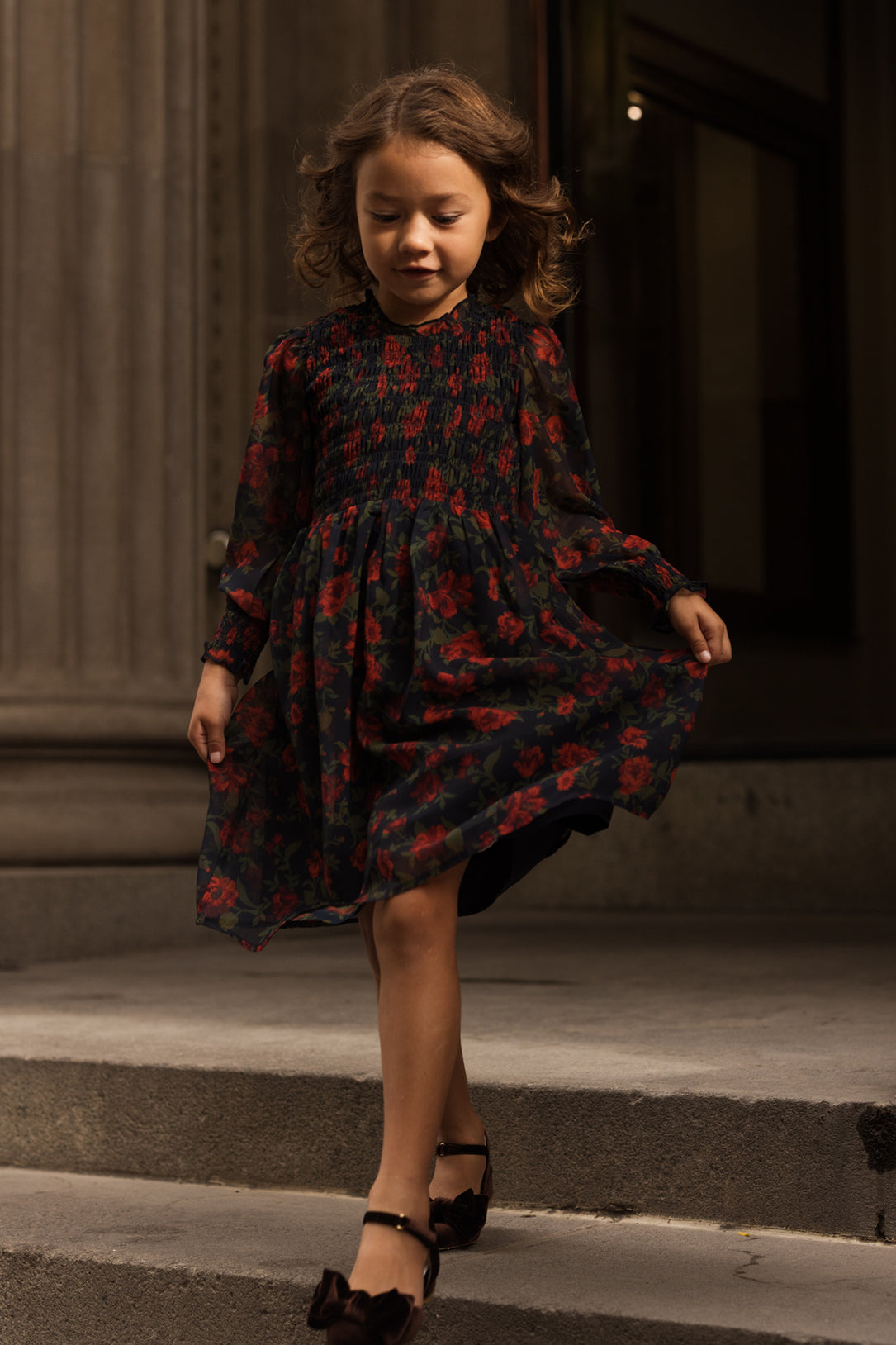 A young girl with curly hair walks down stone steps, holding her skirt of the Ivy City Co Mini Linden Dress in Scarlet Blooms. The dark floral dress features balloon sleeves, and she pairs it with black shoes adorned with bows. Behind her, tall columns frame a grand building entrance.