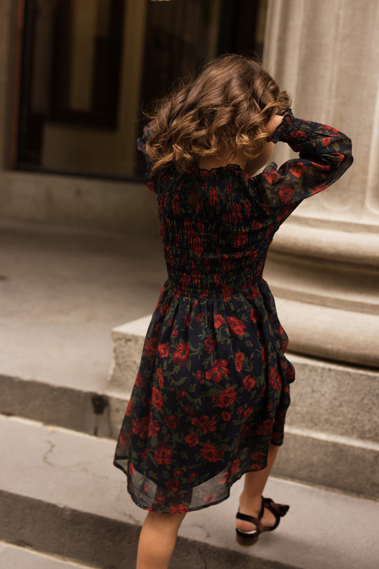 A young girl with curly hair, wearing the Ivy City Co Mini Linden Dress in Scarlet Blooms, walks up stone steps with her hands in her hair. The background features a large column and a building entrance.