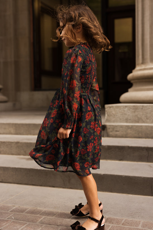 A young girl in the Mini Linden Dress in Scarlet Blooms by Ivy City Co, featuring dark floral fabric and sheer sleeves, stands on stone steps outside a building, her curly hair blowing as she turns slightly, wearing black shoes with bows.