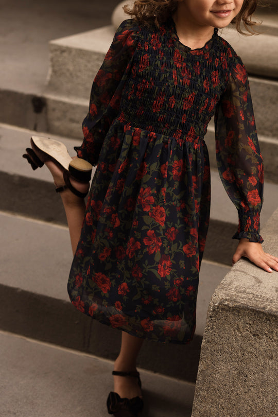 A young girl wearing the Ivy City Co Mini Linden Dress in Scarlet Blooms, a dark long-sleeved dress with a red floral pattern, stands on stone steps, raising one leg behind her while holding her shoe. Her face is mostly out of the frame.