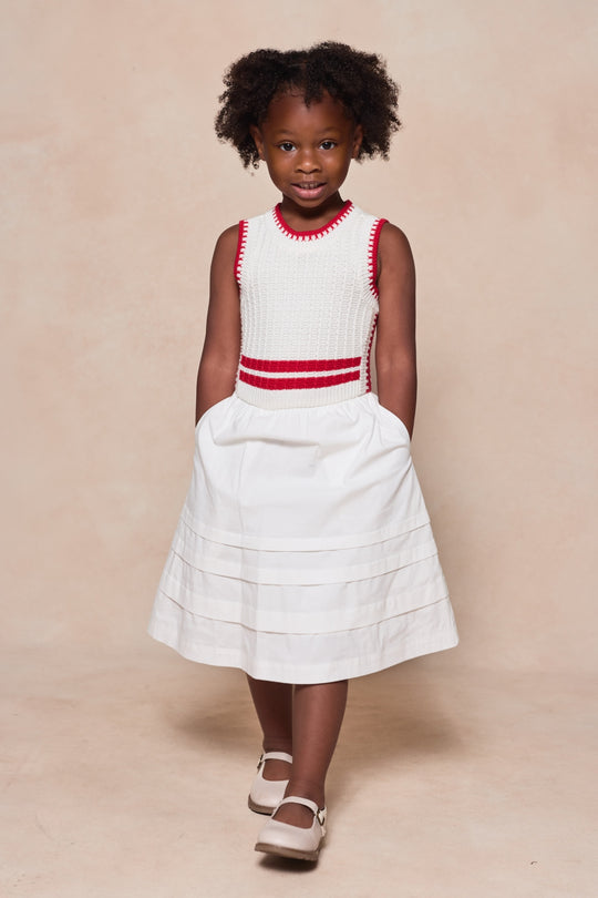 A young girl with curly hair stands with her hands in the pockets of her Ivy City Co Mini Lisbon Dress in Ivory & Firework Red, featuring a sleeveless top and red trim, paired with white shoes against a light beige background.