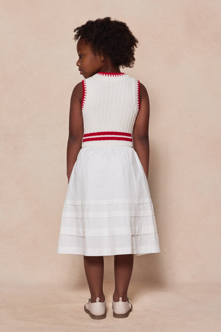 A young girl with curly hair stands facing away, wearing the Ivy City Co Mini Lisbon Dress in Ivory & Firework Red—sleeveless with red trim and stripes near the hem—and white shoes against a beige background. Perfect for Mom and Me matching moments.