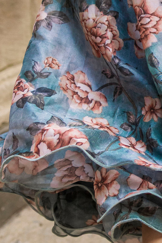 Close-up of the Mini Lisette Dress in Rose Dusk by Ivy City Co, featuring a blue base adorned with a pink floral pattern. The sheer fabric softly reveals ruffled layers underneath, adding texture and movement—making it an elegant option for a fall wedding or a charming choice as a junior bridesmaid dress.
