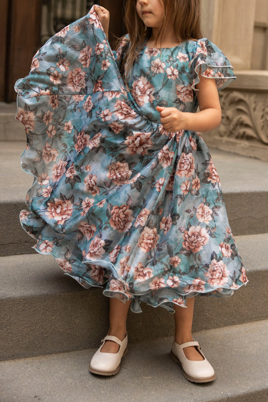 A young girl in Ivy City Co’s Mini Lisette Dress in Rose Dusk and white shoes stands on stone steps, holding out the skirt of her dress with one hand—a charming option for a junior bridesmaid dress at a fall wedding.