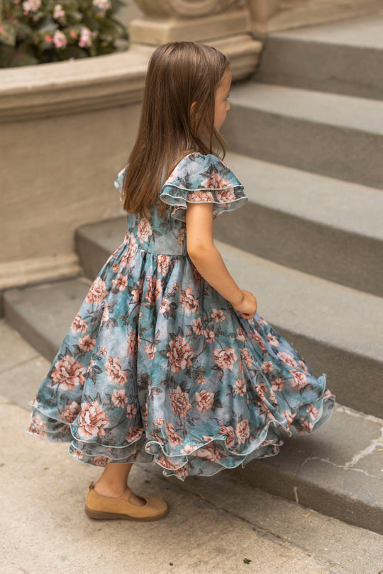 A young girl in the Ivy City Co Mini Lisette Dress in Rose Dusk and tan shoes walks up outdoor stone steps, with flowers and a stone railing in the background.
