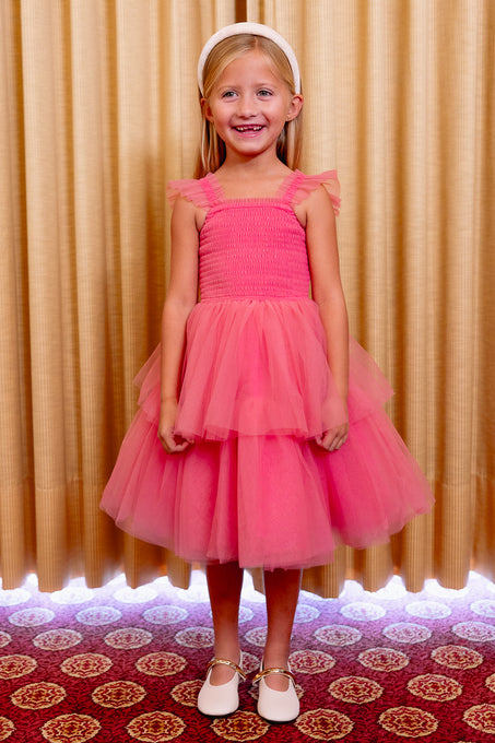 Young girl in a pink dress standing in front of a patterned curtain.