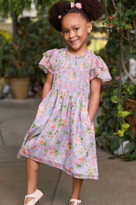 A young girl with puffy hair in two buns and pink flower clips smiles in the Ivy City Co Mini Lottie Dress in Vibrant English Garden, a pastel floral girls’ midi with short ruffled sleeves, posing outdoors among lush green plants.