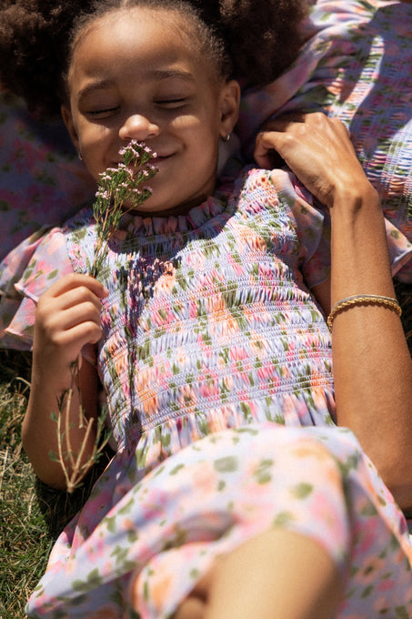 A young girl in an Ivy City Co Mini Lottie Dress in Vibrant English Garden smiles as she lies on the grass holding a flower, next to an adult’s arm with a bracelet—both wearing matching Mini Lottie Dresses.