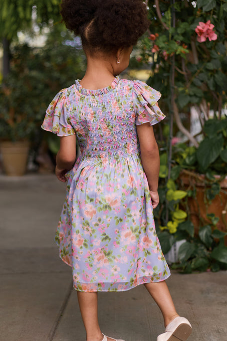 A young girl with curly hair, seen from behind, wears the Mini Lottie Dress in Vibrant English Garden by Ivy City Co and white shoes, standing on a sidewalk surrounded by green plants.