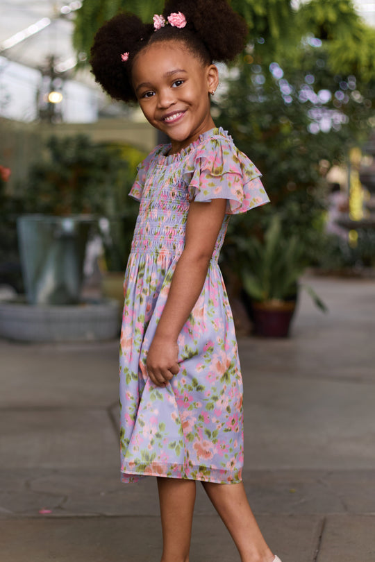 A young girl with natural hair and pink flower clips smiles while wearing the Ivy City Co Mini Lottie Dress in Vibrant English Garden, a pastel floral midi dress with ruffled sleeves, as she stands sideways in a greenhouse filled with green plants.