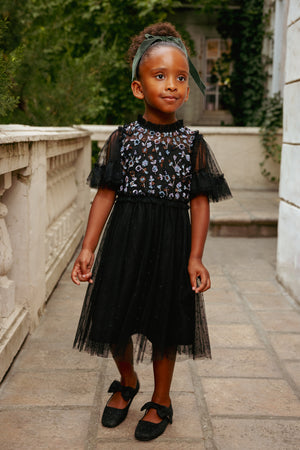 Young girl in a black dress with floral embroidery standing outdoors.
