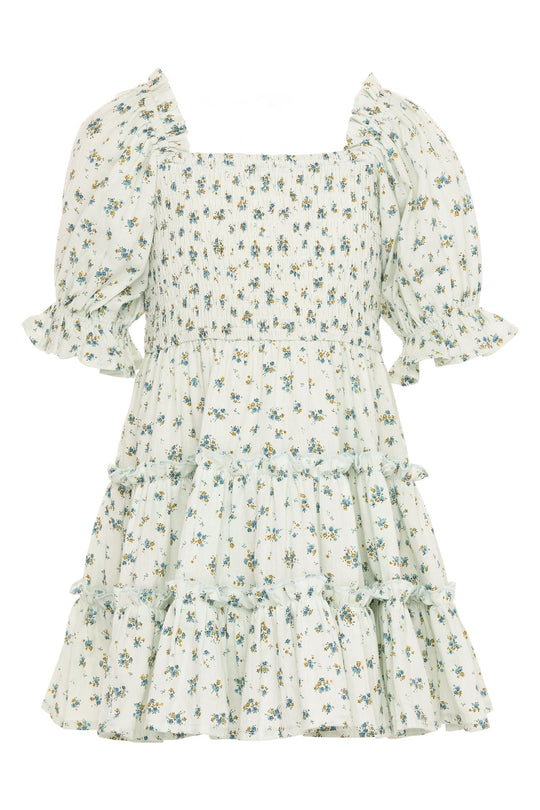 The Mini Madeline Dress in Blue by Ivy City Co is a light-colored, short-sleeved girls’ cotton dress with a square neckline, smocked bodice, puffed sleeves, and a ruffled tiered skirt. It features an allover small blue floral print—making it a charming choice for any occasion.