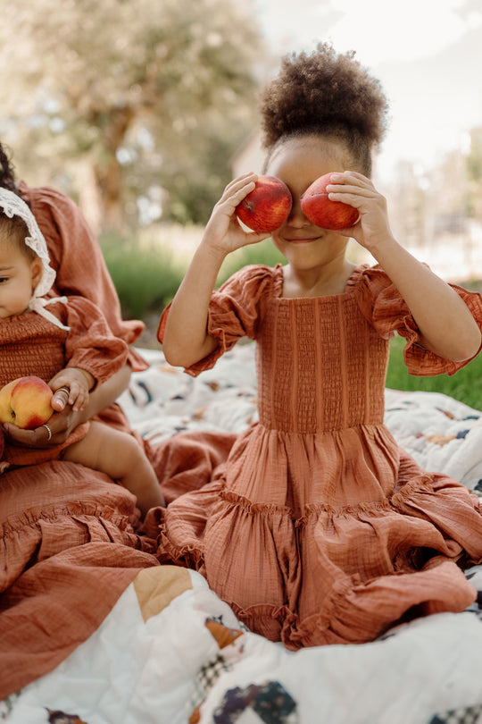 A young girl wears the Ivy City Co Mini Madeline Dress in Apple Cider, featuring puff sleeves, as she sits on a blanket smiling and holds two apples over her eyes. Beside her, a baby in a matching dress with a smocked bodice holds an apple.