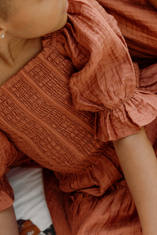 A child wearing the Ivy City Co Mini Madeline Dress in Apple Cider, featuring a textured fabric, smocked bodice, and puff sleeves, sits close to another person dressed similarly, both partially visible.