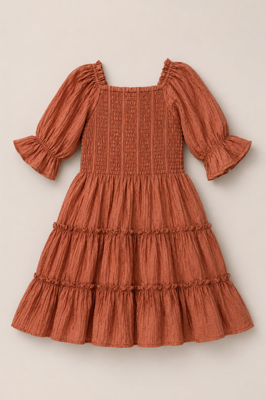 The Mini Madeline Dress in Apple Cider by Ivy City Co features a warm, apple cider hue with long puff sleeves, ruffled cuffs, a smocked bodice, and a tiered knee-length skirt, shown laid flat on a neutral background.