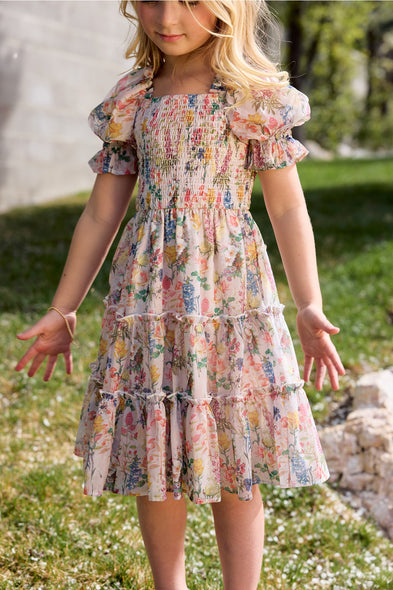 A young girl with long blonde hair wears the Ivy City Co Mini Madeline Dress in Cordelia Floral—a light floral birthday dress with puffed sleeves and a smocked bodice—while standing on grass near a stone wall in sunlight.