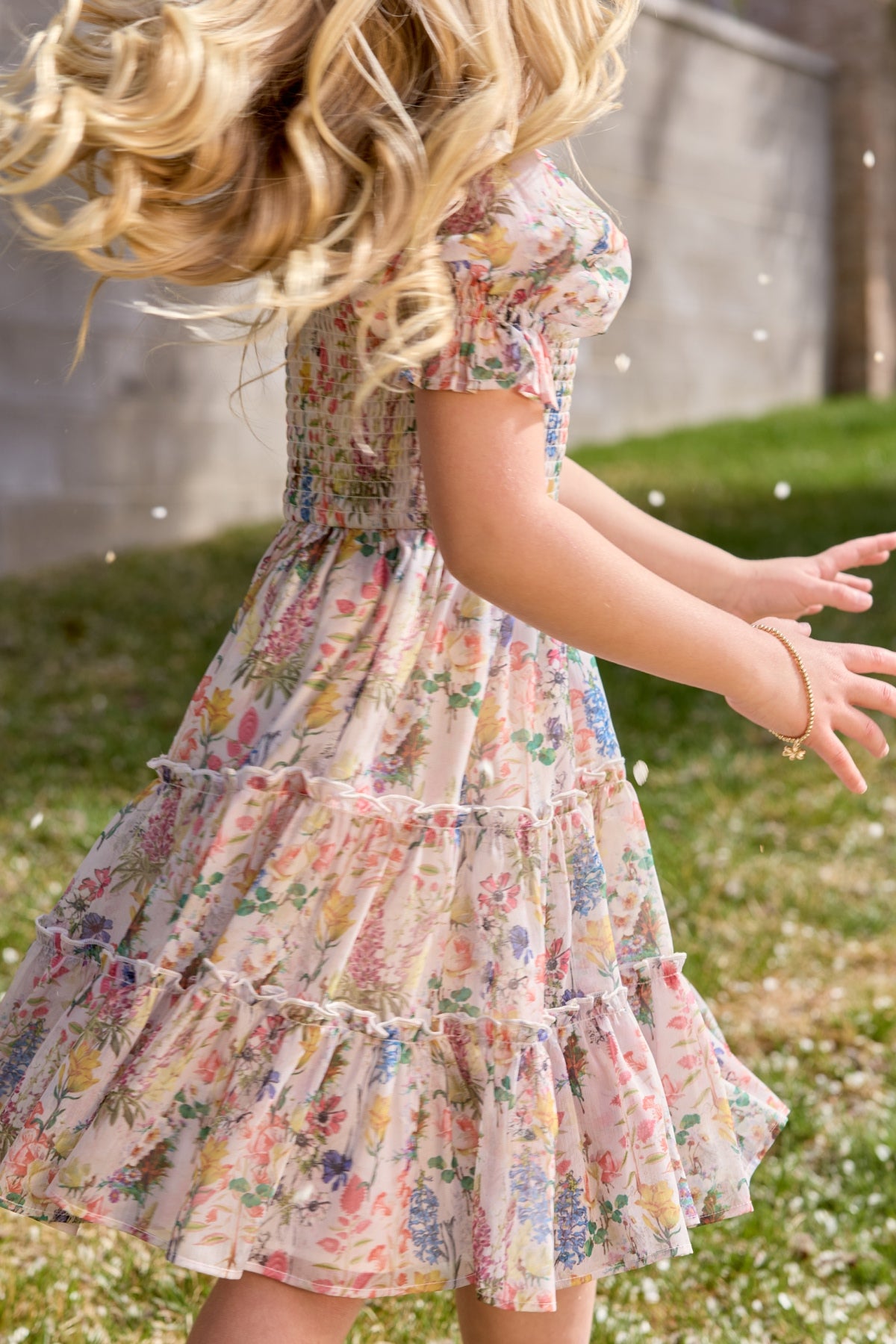 A young girl with long, wavy blonde hair twirls outside in the Ivy City Co Mini Madeline Dress in Cordelia Floral. Sunlight catches her flowing hair and skirt as she stands on grass by a concrete wall with white petals swirling around her.