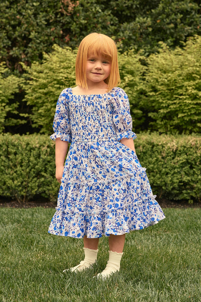 A young girl wears the Ivy City Co Mini Madeline Dress in Ditsy Cobalt Floral, paired with white socks and cream shoes, standing on grass with lush greenery—ideal for a charming Mom and Me matching moment.
