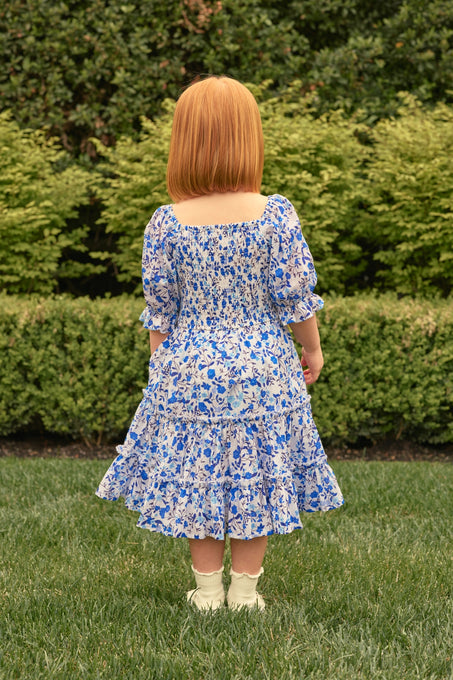A young girl with straight red hair wears the Ivy City Co Mini Madeline Dress in Ditsy Cobalt Floral, paired with white socks and shoes, standing on grass and facing away from the camera, with green bushes in the background.