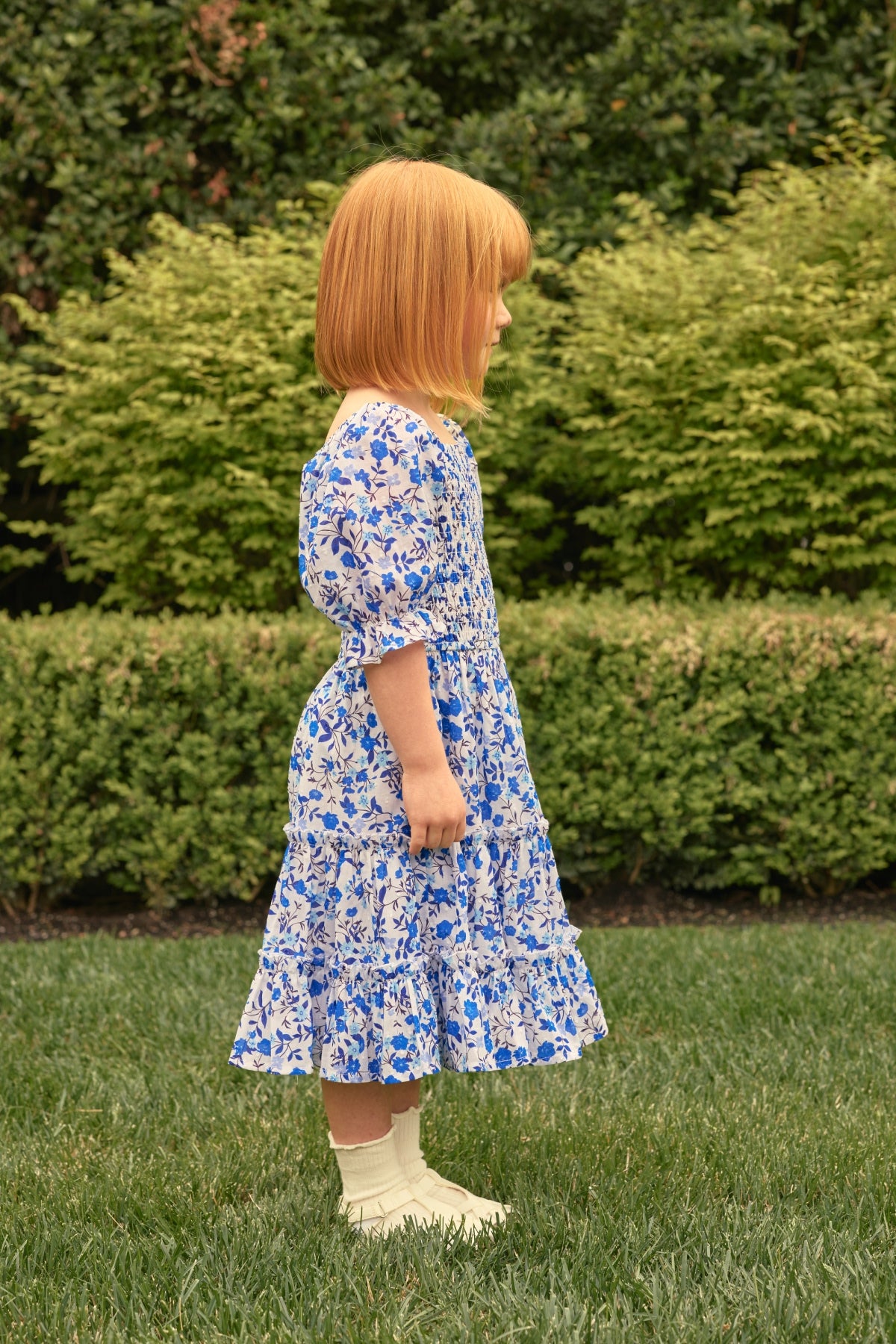A young child with straight red hair, wearing the Ivy City Co Mini Madeline Dress in Ditsy Cobalt Floral and white socks, stands in profile on a grassy lawn with green bushes in the background.