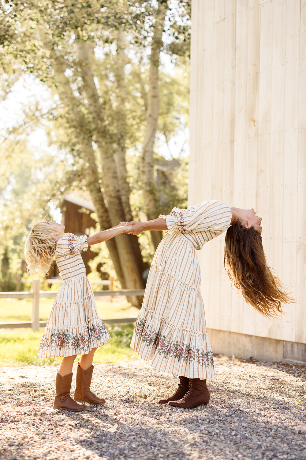 A woman and a girl, both wearing Ivy City Co’s Mini Madeline Dress in Embroidered Cider Grid with smocked bodices and boots, hold hands outdoors near a wooden building as sunlight filters through the trees; the woman leans back dramatically while the girl pulls forward, both smiling.