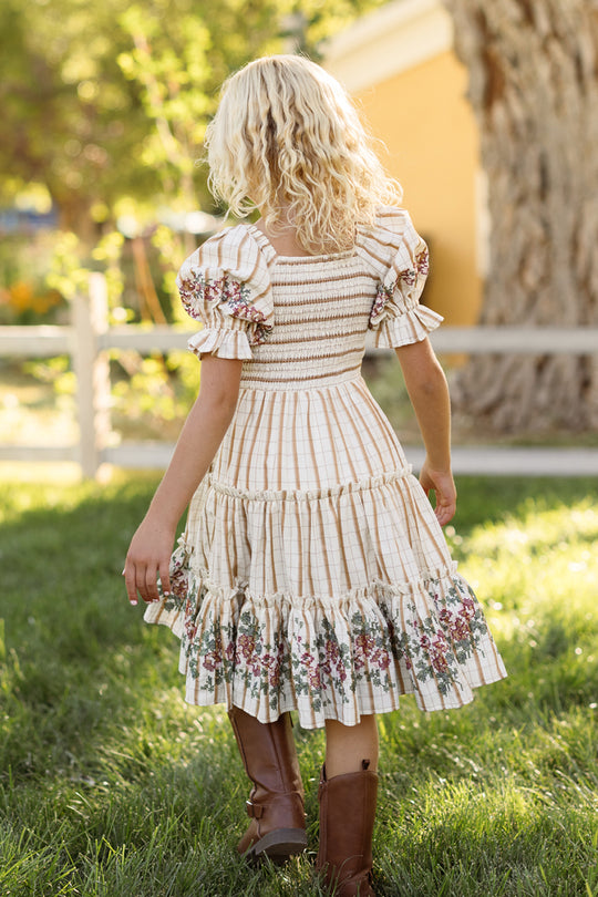 A young girl with curly blonde hair, dressed in the Ivy City Co Mini Madeline Dress in Embroidered Cider Grid with a smocked bodice and ruffled floral details, pairs the look with brown boots as she walks on green grass near a wooden fence and large tree on a sunny day.