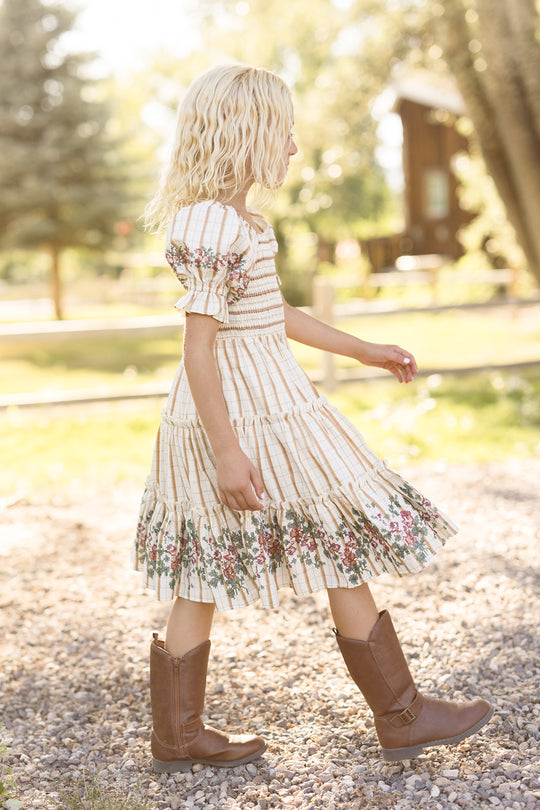 A young girl with blonde hair walks outside on a gravel path, wearing the Ivy City Co Mini Madeline Dress in Embroidered Cider Grid, which features a smocked bodice. She pairs the floral dress with brown boots, while trees and a wooden building appear in the sunny background.