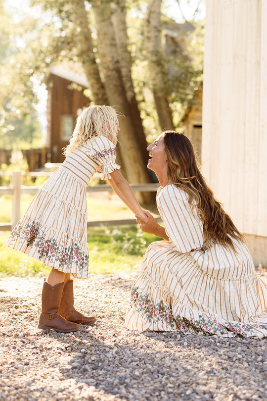 A woman kneels and smiles at a young girl, both wearing matching Ivy City Co Mini Madeline Dresses in Embroidered Cider Grid. The girl’s dress features a charming smocked bodice, which she pairs with brown boots. Sunlight filters through the trees in the background, with a wooden building visible.