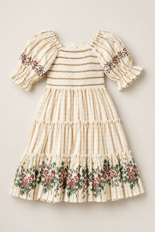 The Mini Madeline Dress in Embroidered Cider Grid by Ivy City Co is a cream-colored, plaid girls’ dress with a smocked bodice, puffed sleeves, and ruffled hem. Charming floral embroidery in green and burgundy decorates the sleeves and skirt.