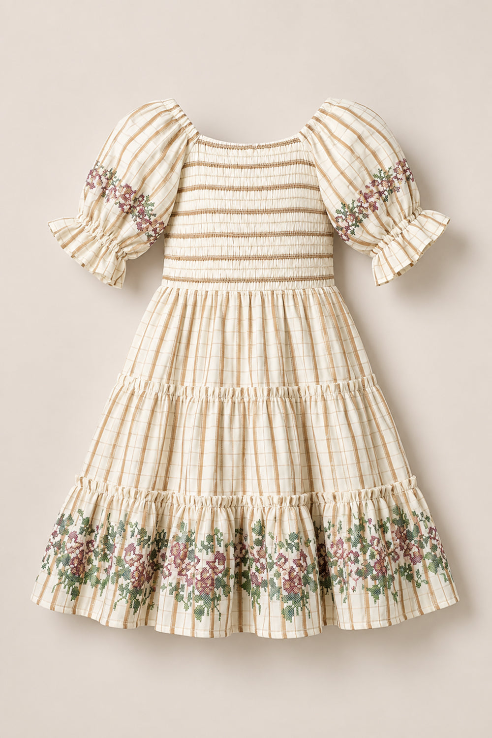 The Mini Madeline Dress in Embroidered Cider Grid by Ivy City Co is a cream plaid girls’ dress featuring puffed short sleeves, a smocked bodice, and a ruffled hem. Delicate floral embroidery adorns the sleeves and bottom ruffle. The dress is displayed laid flat on a neutral background.