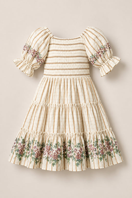 The Mini Madeline Dress in Embroidered Cider Grid by Ivy City Co is a cream plaid girls’ dress featuring puffed short sleeves, a smocked bodice, and a ruffled hem. Delicate floral embroidery adorns the sleeves and bottom ruffle. The dress is displayed laid flat on a neutral background.