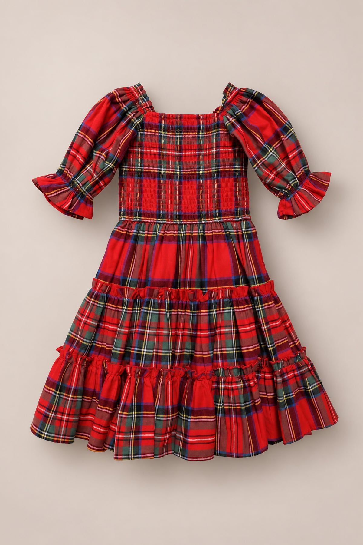 The Mini Madeline Dress in Holiday Plaid by Ivy City Co features puffed sleeves, ruffle cuffs, a smocked bodice, and a tiered ruffle skirt. Shown laid flat on a beige background—this festive girls’ midi dress is perfect for the holidays.