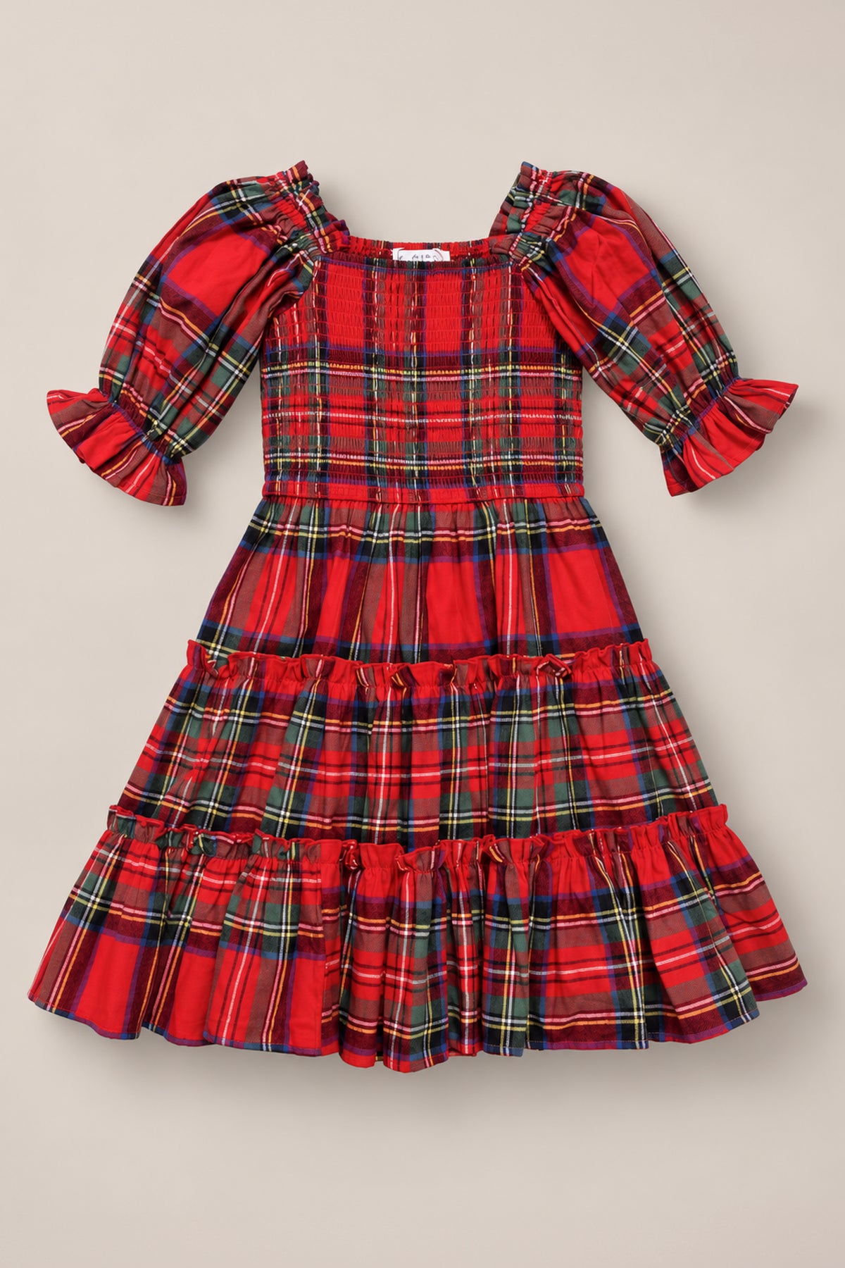 The Mini Madeline Dress in Holiday Plaid by Ivy City Co is a festive midi dress for girls crafted from red plaid cotton. It features a smocked bodice, puffed half sleeves with ruffled cuffs, and a tiered, ruffled skirt. The classic holiday plaid pattern is accented with green, blue, and yellow lines for a charming look.
