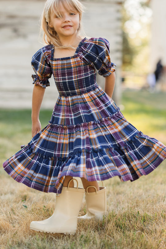 A young girl in the Ivy City Co Mini Madeline Dress in Midnight Plaid, featuring a smocked bodice, twirls on the grass with a smile, wearing cream rain boots. The background is softly blurred with greenery and part of a white building visible.