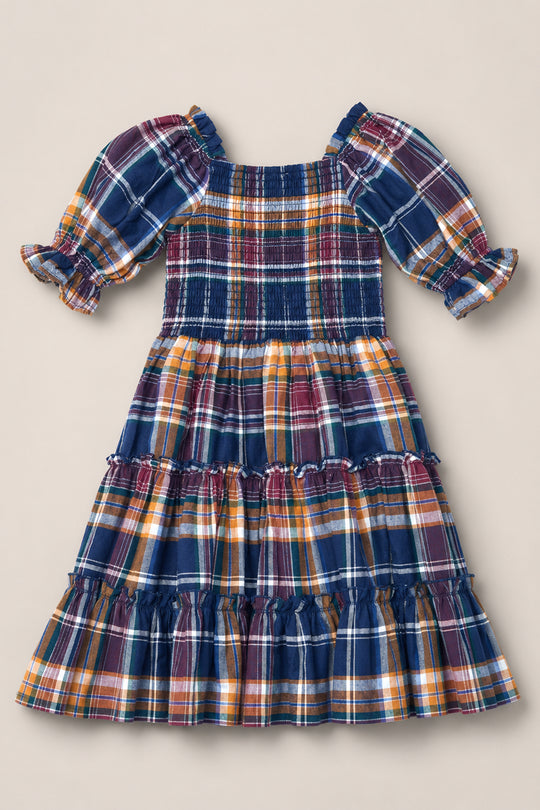 The Mini Madeline Dress in Midnight Plaid by Ivy City Co is a girls’ knee-length dress featuring ruffled tiers, puff sleeves, and a smocked bodice. Crafted from soft fabric, it showcases a multicolored plaid pattern in blue, yellow, and red tones.