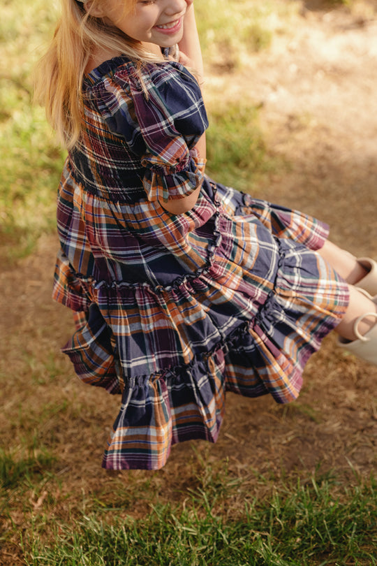 A young girl sits smiling on a swing, sunlight casting shadows over the grass beneath her, wearing Ivy City Co’s Mini Madeline Dress in Midnight Plaid, featuring puff sleeves and a smocked bodice.
