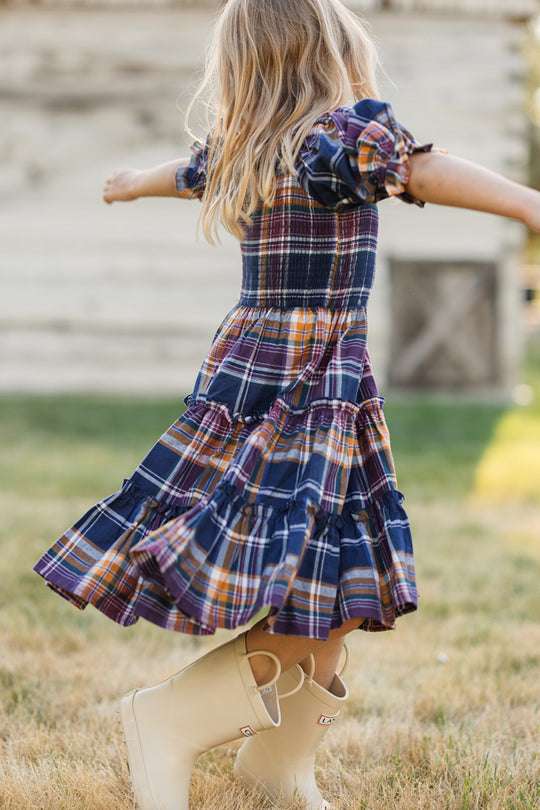A young girl with blonde hair twirls outside on the grass, wearing the Ivy City Co Mini Madeline Dress in Midnight Plaid, which features a smocked bodice and ruffles, paired with light-colored rain boots. A blurred wooden structure appears in the background.