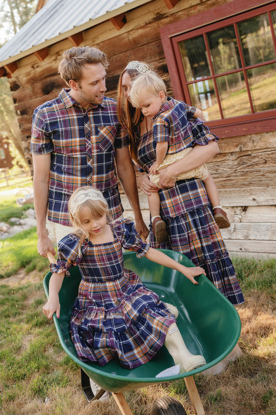 A family in matching plaid outfits smiles outside a log cabin. The mother, dressed in Ivy City Co’s Mini Madeline Dress in Midnight Plaid—with its puff sleeves and smocked bodice—holds a toddler while the father pushes a young girl in a green wheelbarrow. Sunlight filters through the trees in the background.