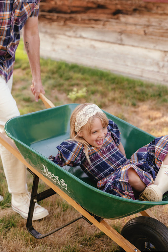 A young child wearing the Ivy City Co Mini Madeline Dress in Midnight Plaid with puff sleeves and a lace headband smiles while sitting in a green wheelbarrow, being pushed by an adult outdoors near a wooden building.