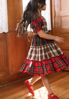 A young girl with long hair twirls indoors on a wooden floor near wood-paneled walls and a window with lace curtains, wearing the Mini Madeline Dress in Mixed Plaid by Ivy City Co and red shoes.
