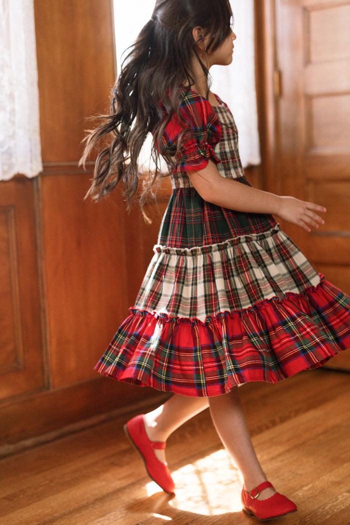 A young girl with long hair twirls indoors on a wooden floor near wood-paneled walls and a window with lace curtains, wearing the Mini Madeline Dress in Mixed Plaid by Ivy City Co and red shoes.