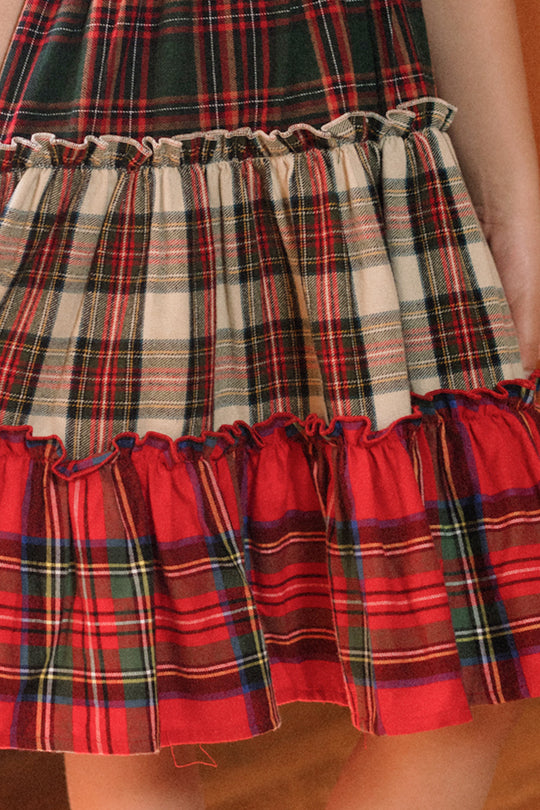 A close-up of a person wearing the Ivy City Co Mini Madeline Dress in Mixed Plaid, featuring layered red, green, and white plaid patterns and ruffled edges, with their hand resting by their side.