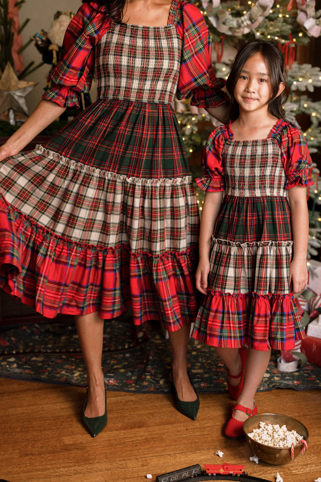 A woman in a Madeline Dress and a girl in the Mini Madeline Dress in Mixed Plaid by Ivy City Co stand by a decorated Christmas tree. The woman holds out her skirt while the girl smiles happily in her plaid dress. At their feet, a toy train and scattered popcorn add to the festive scene.