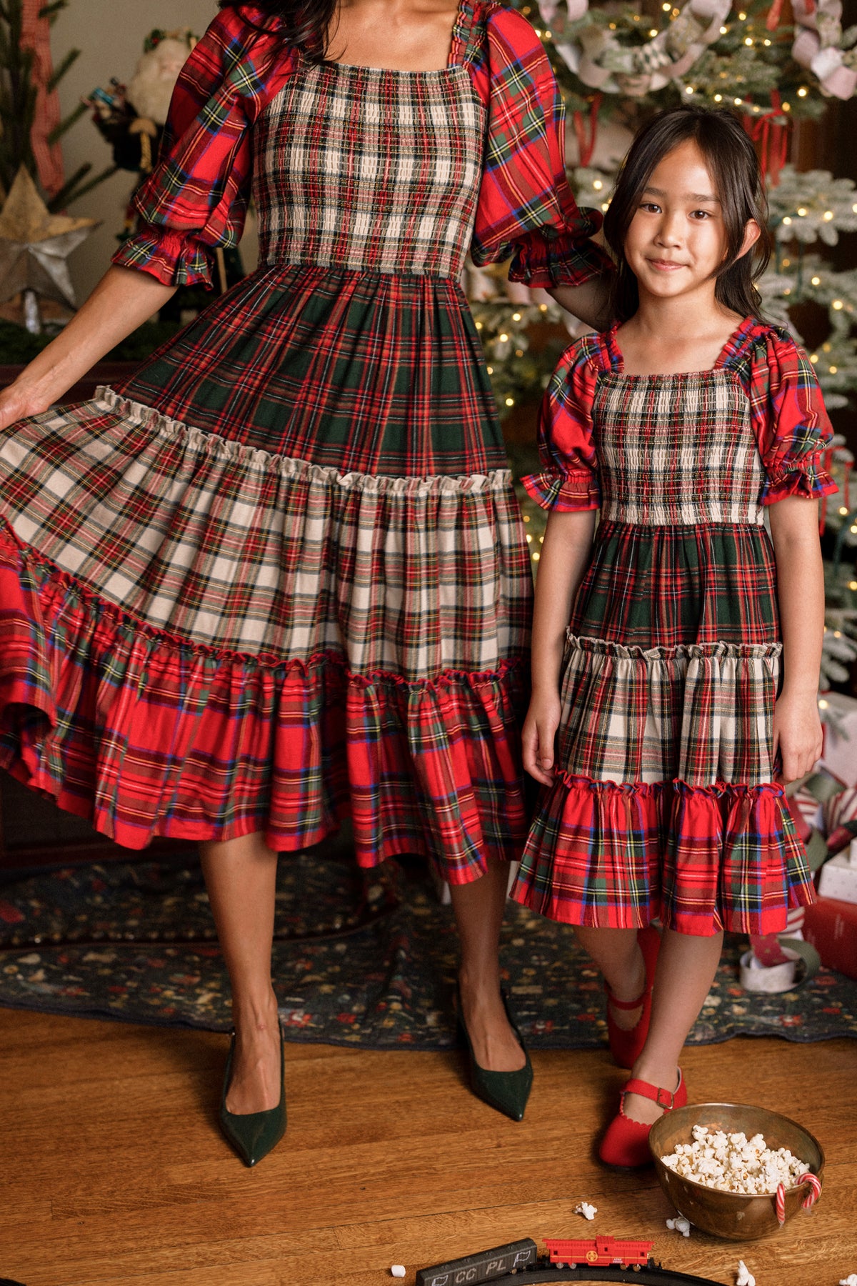 A woman in a Madeline Dress and a girl in the Mini Madeline Dress in Mixed Plaid by Ivy City Co stand by a decorated Christmas tree. The woman holds out her skirt while the girl smiles happily in her plaid dress. At their feet, a toy train and scattered popcorn add to the festive scene.