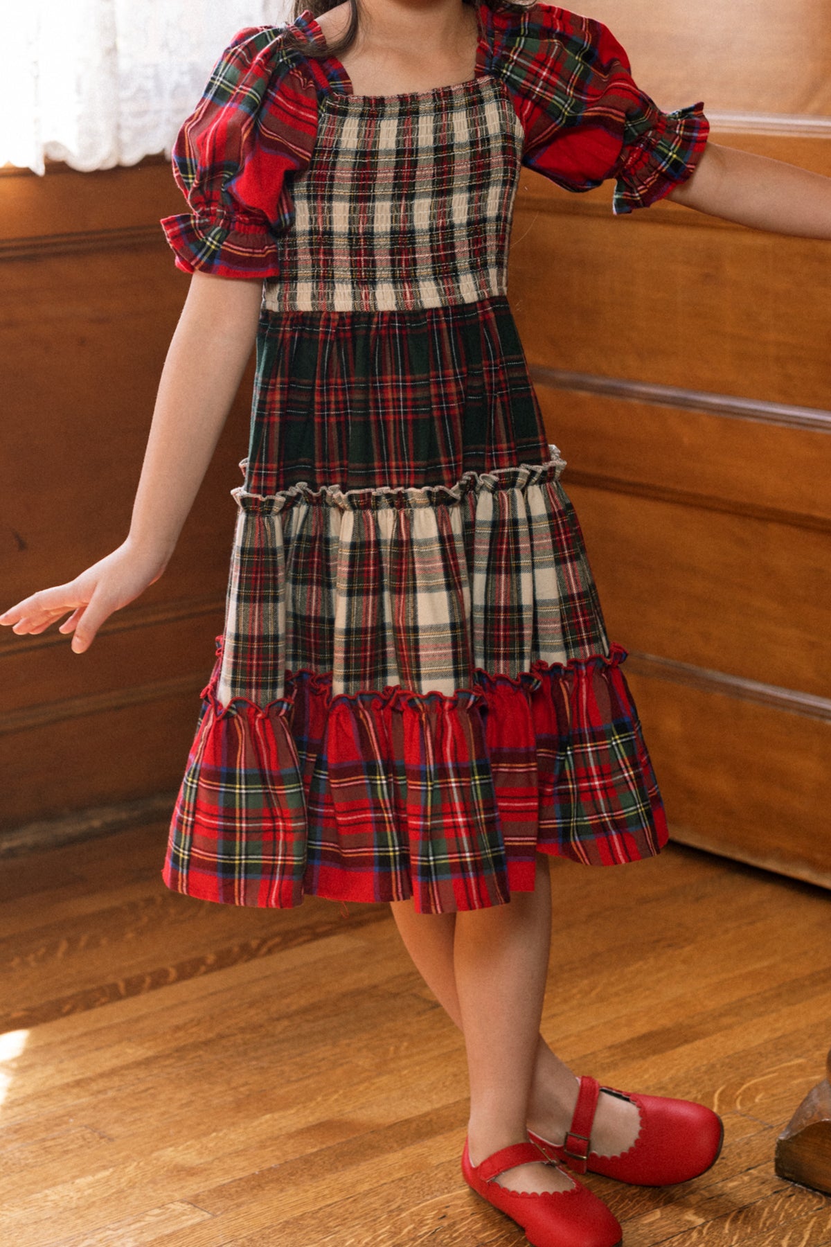 A child wearing the Mini Madeline Dress in Mixed Plaid by Ivy City Co—a delightful girls’ holiday dress with puffed sleeves and ruffle details in classic red and green plaid—stands on a wooden floor in a warmly lit room, her red shoes finishing the festive look for a perfect Christmas ensemble.